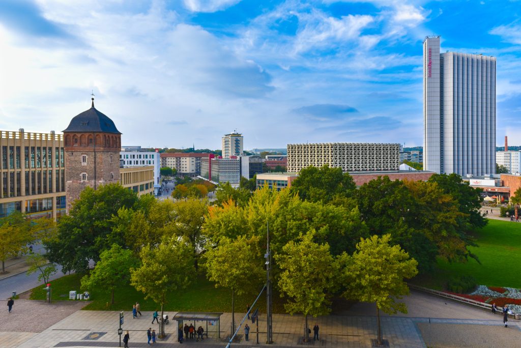 Das Bild zeigt eine Luftaufnahme von der Chemnitzer Innenstadt. Im Mittelpunkt steht eine grüne Parkanlage vor blauem Himmel. Die Parkanlage wird eingerahmt von verschiedenen modernen Hochhäusern und einem etwas älter aussehenden Turm.