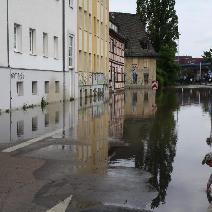 Das Bild zeigt eine stark versiegelte Straße mit Wohngebäuden in Halle, die vom Hochwasser betroffen ist. Das Wasser steht ca. 20 Zentimeter auf der Straße.