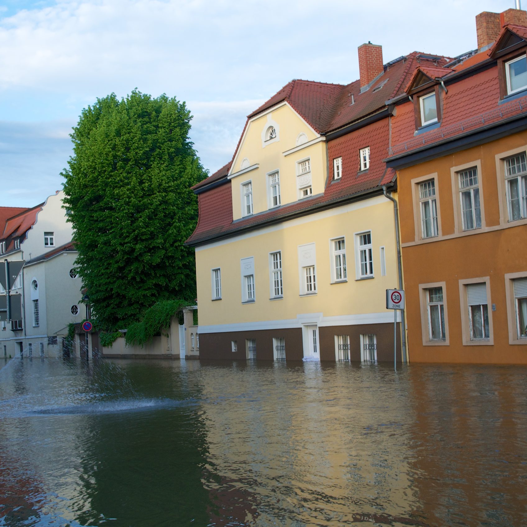 Das Bild zeigt eine überflutete Straße während des Hochwassers in Halle 2013. Das Wasser steht ca. einen halben Meter hoch und verdeckt die Straße komplett. Im Hintergrund steht eine Häuserreihe und ein Baum.